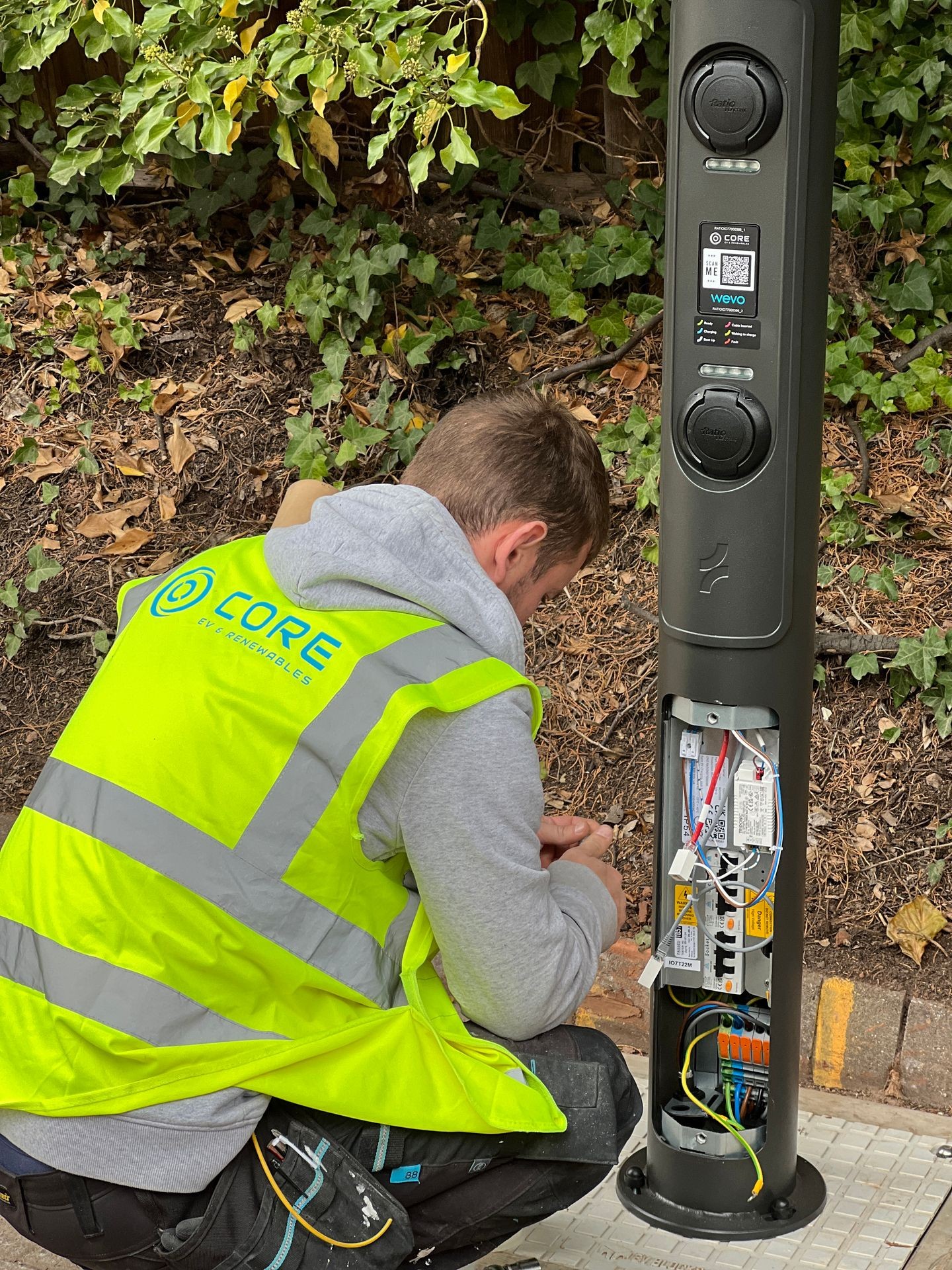 Worker in hi-vis vest installing cables in an outdoor charging station by a leafy area.