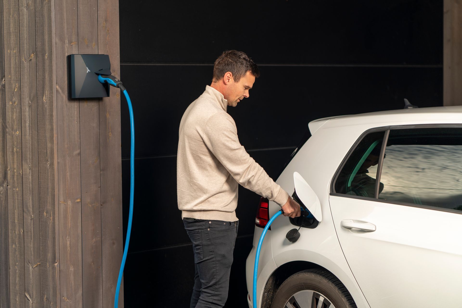 Person charging an electric car at a wall-mounted charging station with a blue cable.