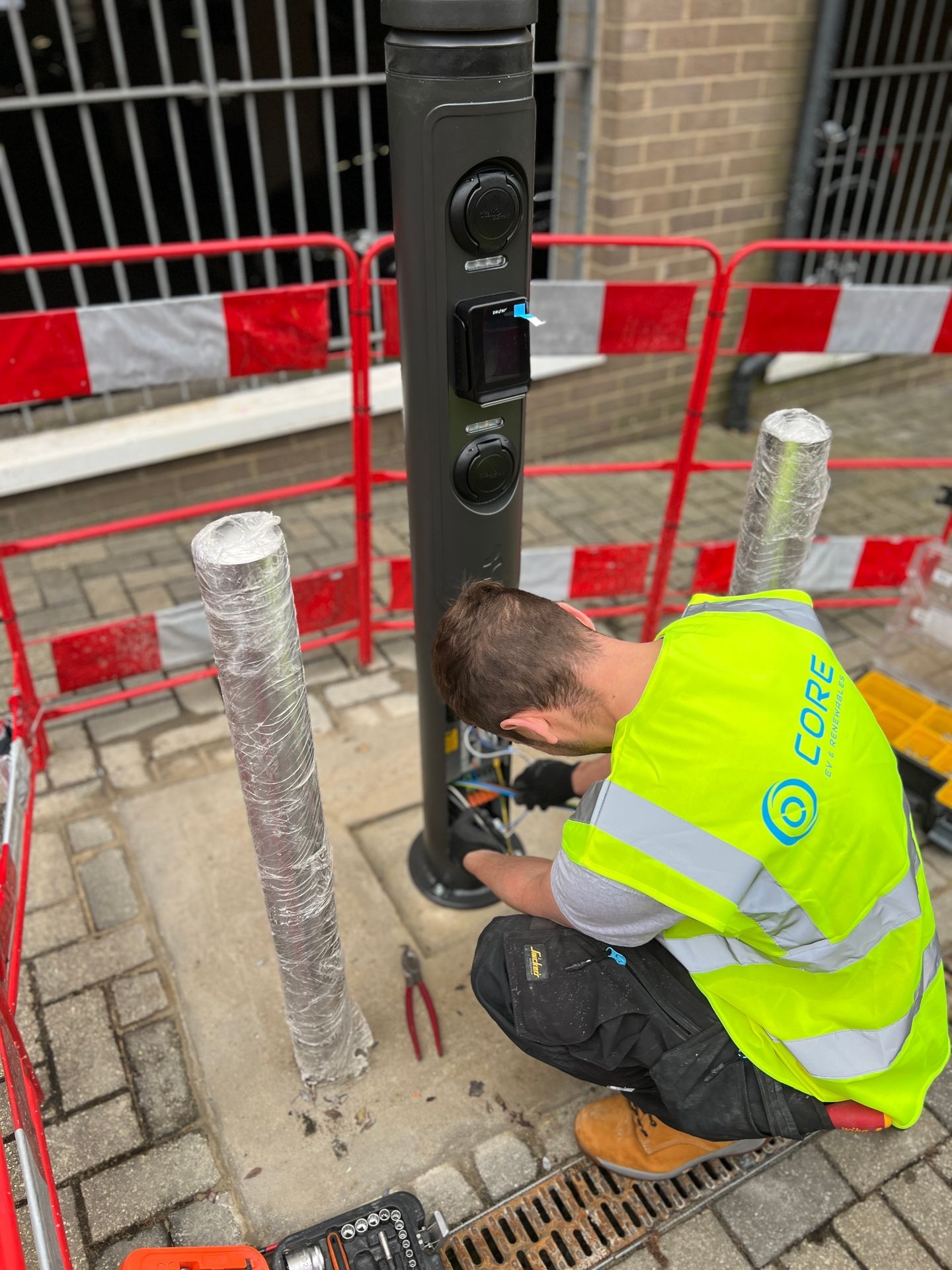 Worker in hi-vis vest installing components on a pole beside a red barrier.