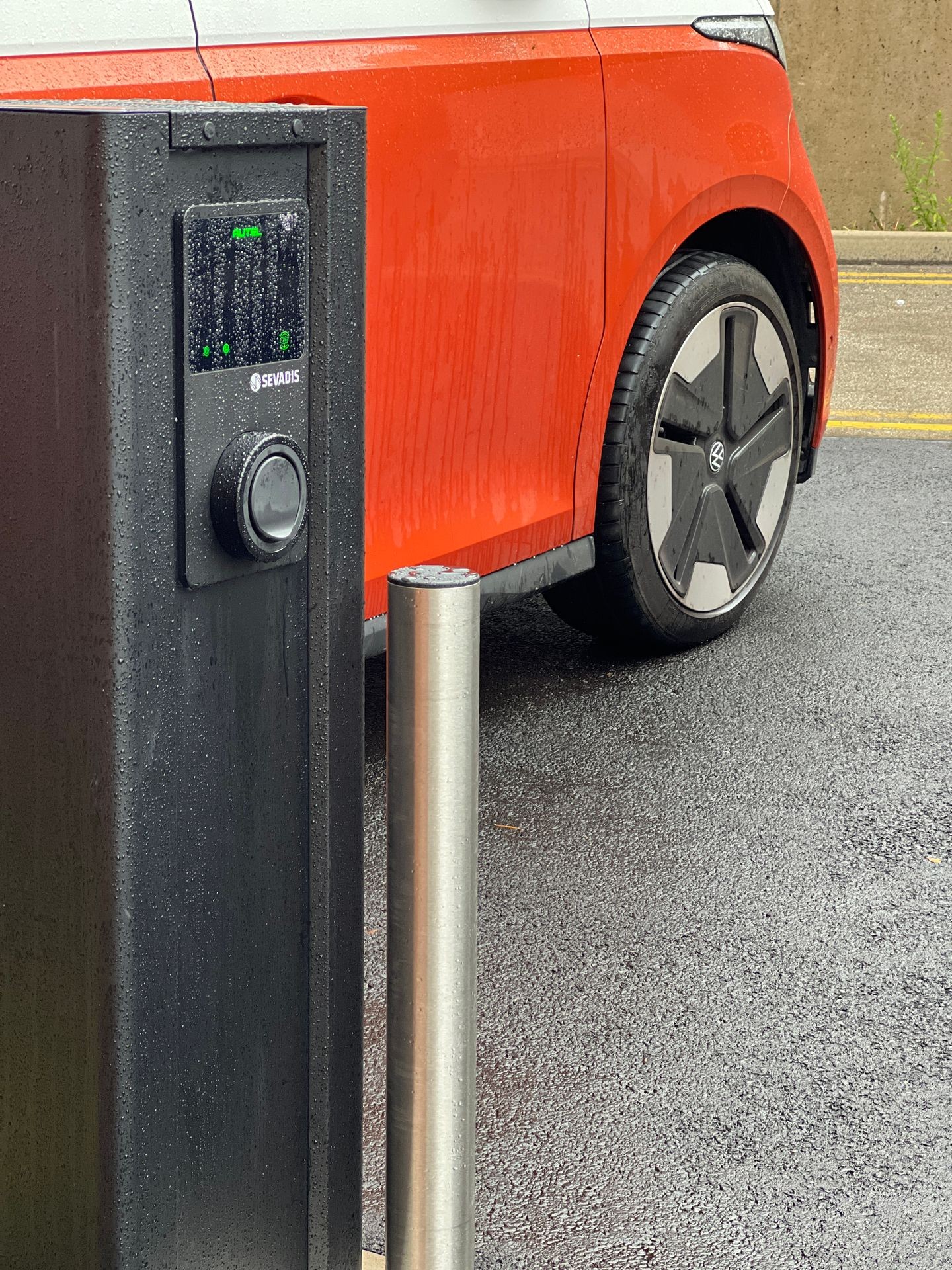 Red vehicle parked next to a wet electric vehicle charging station on a rainy day.